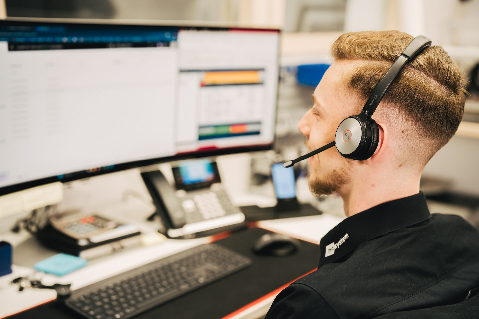 Marvin Böhmler von hcsystem telefoniert mit Headset am Arbeitsplatz, daneben ein IP-Tischtelefon und ein Mobilteil.